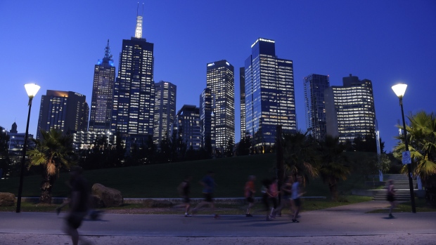 Evening joggers run through Birrarung Marr park as commercial buildings stand illuminated in the background in Melbourne, Australia, on Friday, April 28, 2017. Australia has some of the world's largest reserves of fossil fuels but is running short on gas. That's raised the prospect of widespread power outages and stoked a national debate over energy policy. 