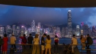 People stand along Victoria Harbour in the Tsim Sha Tsui district as Two International Finance Center (IFC), center right, Bank of China Tower, center left, and other buildings on Hong Kong island stand illuminated at dusk in Hong Kong, China, on Monday, April 29, 2019. Hong Kong's gross domestic product will expand at a slower pace in the first quarter than the fourth because of the high base in the first three months of last year and due to external uncertainties, according to the city's financial secretary on April 28. 