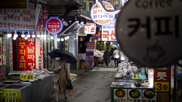 A pedestrian wearing a protective mask and holding an umbrella walks past stores at Namdaemun Market in Seoul, South Korea, on Friday, Feb. 28, 2020. The South Korean government announced a raft of subsidies and consumer incentives to support an economy hit hard by the spread of coronavirus. Photographer: SeongJoon Cho/Bloomberg