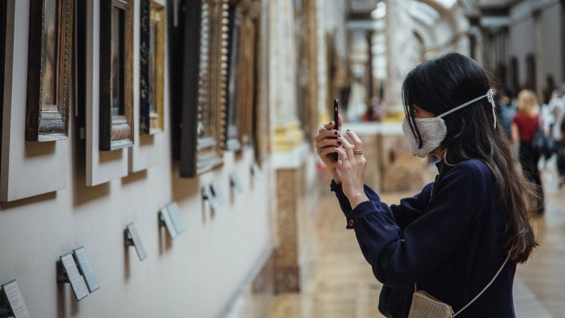 A visitor wearing a protective face mask takes a smartphone photograph of paintings hanging in a gallery in the reopened Louvre Museum in Paris, France, on Monday, July 6, 2020. The Louvre had 9.6 million visitors last year.