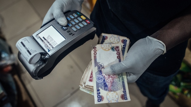 A vendor wearing protective latex gloves holds Nigerian naira banknotes and a contactless payment terminal in a store in Lugbe district in Abuja, Nigeria, on Wednesday, June 3, 2020. The government of Nigeria, whose revenue could be slashed by more than half this year due to the oil-price slump, finalized plans for a revised budget that keeps spending almost intact, and that will mean more borrowing. Photographer: KC Nwakalor/Bloomberg
