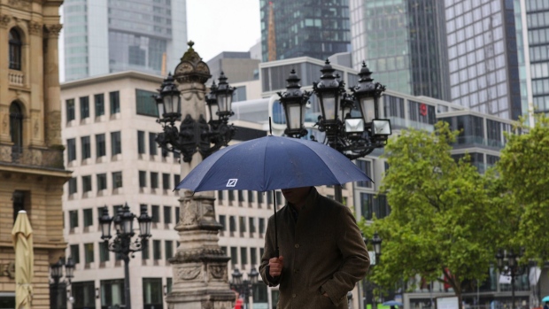 A city worker with a Deutsche Bank AG branded umbrella near skyscrapers and office properties in the financial district in Frankfurt, Germany, on Wednesday, May 26, 2021. Europe’s labor market may recover more slowly from the pandemic than its economy, according to a study by Accenture.