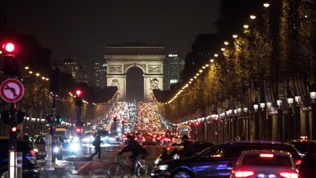 Vehicles sit at traffic lights as the Arc de Triomphe stands beyond at night in Paris.