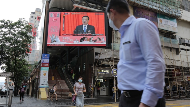 Pedestrians wearing protective masks walk past a screen playing a news report on Chinese Premier Li Keqiang speaking at the National People's Congress in Hong Kong, China, on Friday, May 22, 2020. Democracy advocates called for protests against sweeping national security legislation China introduced Friday, as authorities in Beijing vowed to end what they called a "defenseless" posture due to "those trying to sow trouble."
